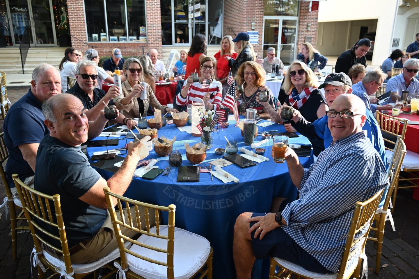 A group of people sit around a table outdoors, smiling and raising drinks at a festive gathering.