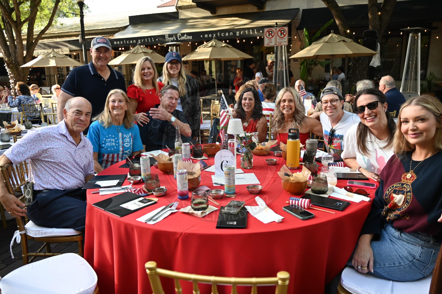 A group of people smile around a red table at an outdoor restaurant decorated with American decor.