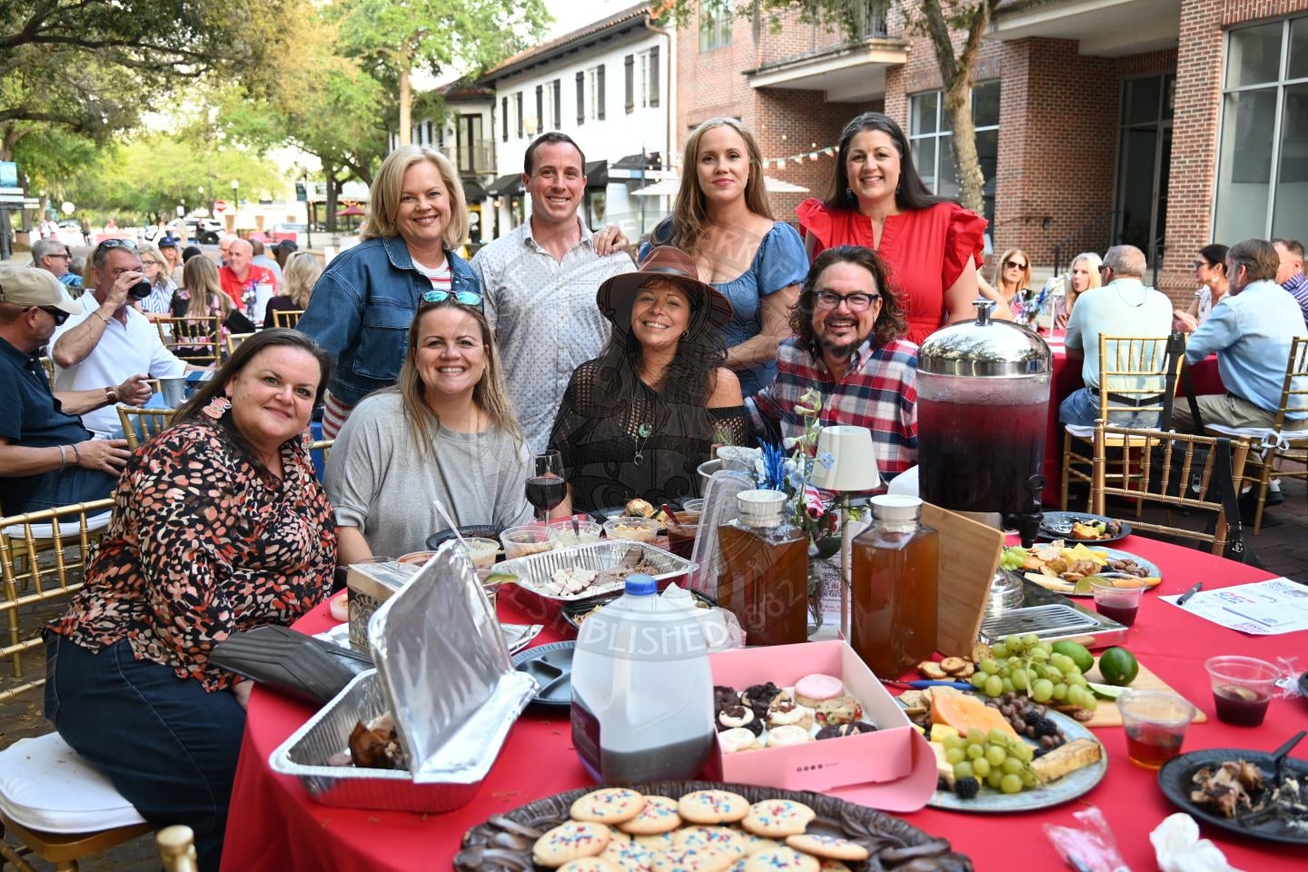 A group of people smile at an outdoor event, gathered around a table with food, drinks, and desserts.