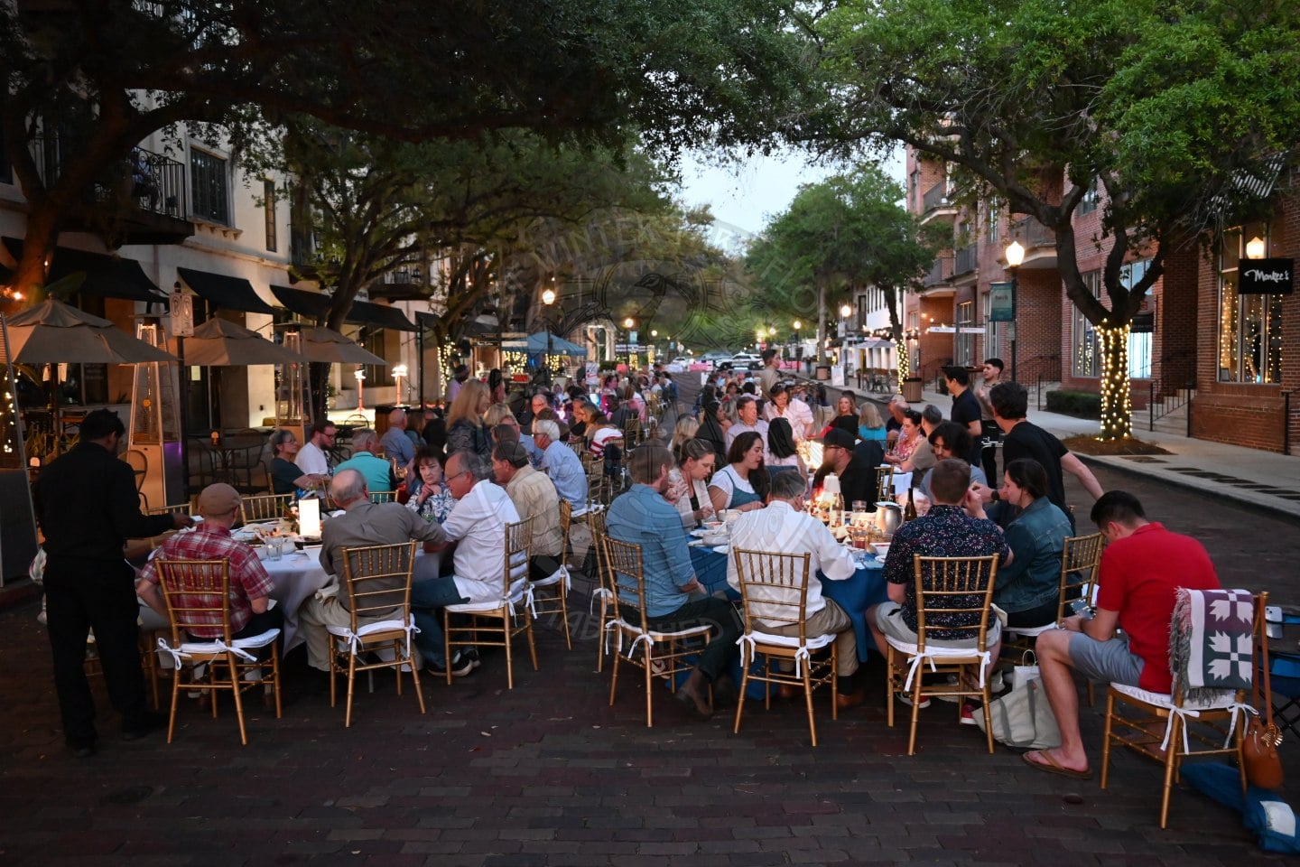 A large group dines at long tables set up on a tree-lined city street in the evening, with twinkling lights and candles.
