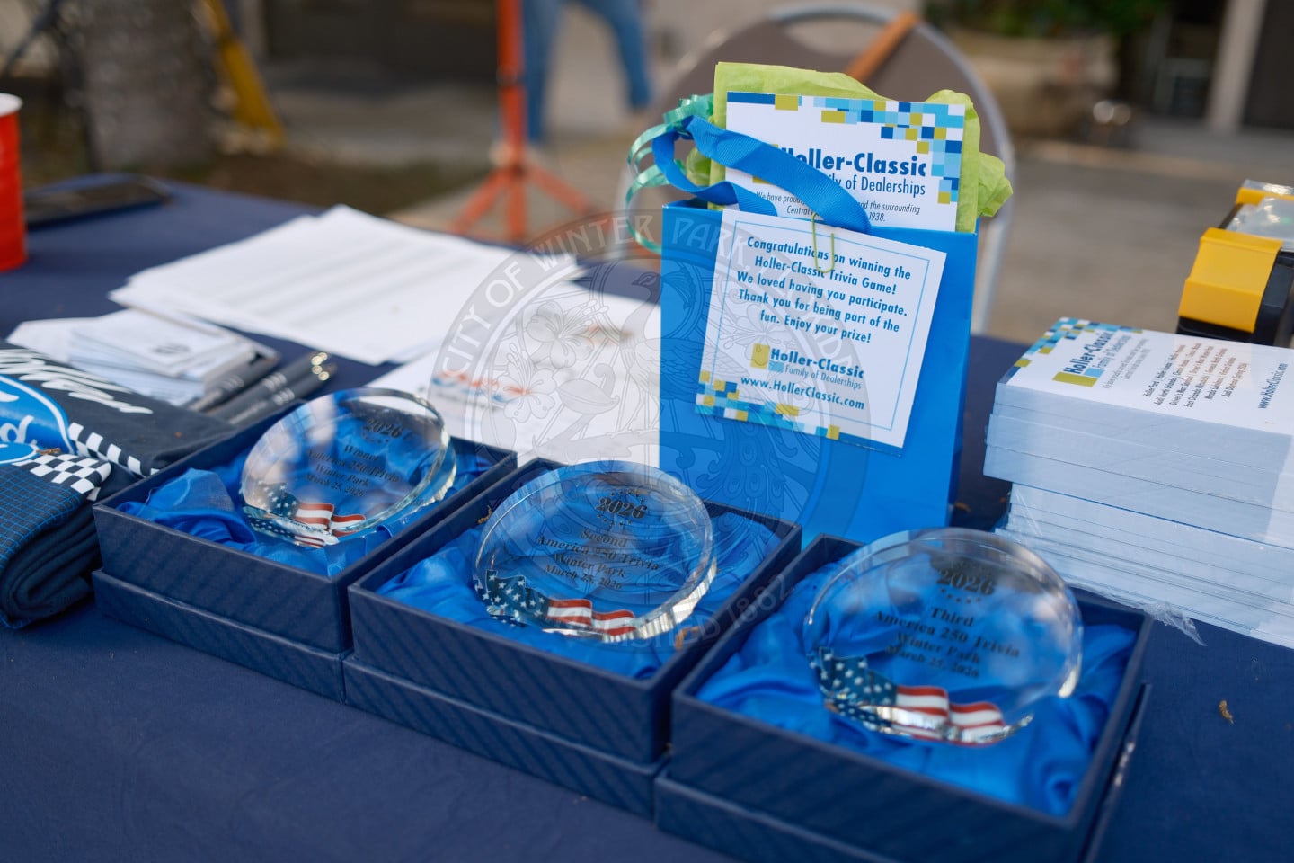 Three glass awards with American flags inside are displayed on a table next to event flyers and informational papers.