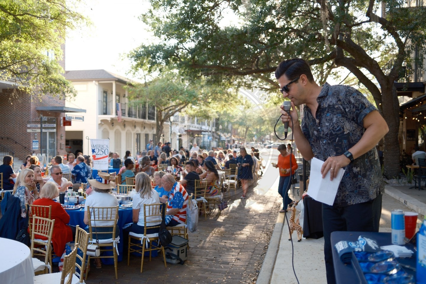 A man speaks into a microphone to a large outdoor crowd seated at tables on a tree-lined street during the day.