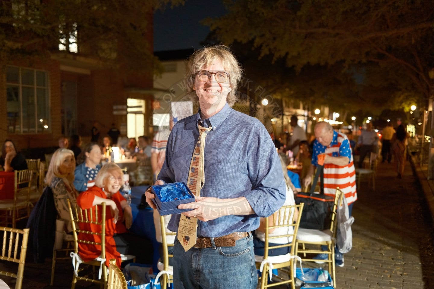A smiling man in glasses holds a blue glass trophy at an outdoor evening party with people seated at decorated tables.