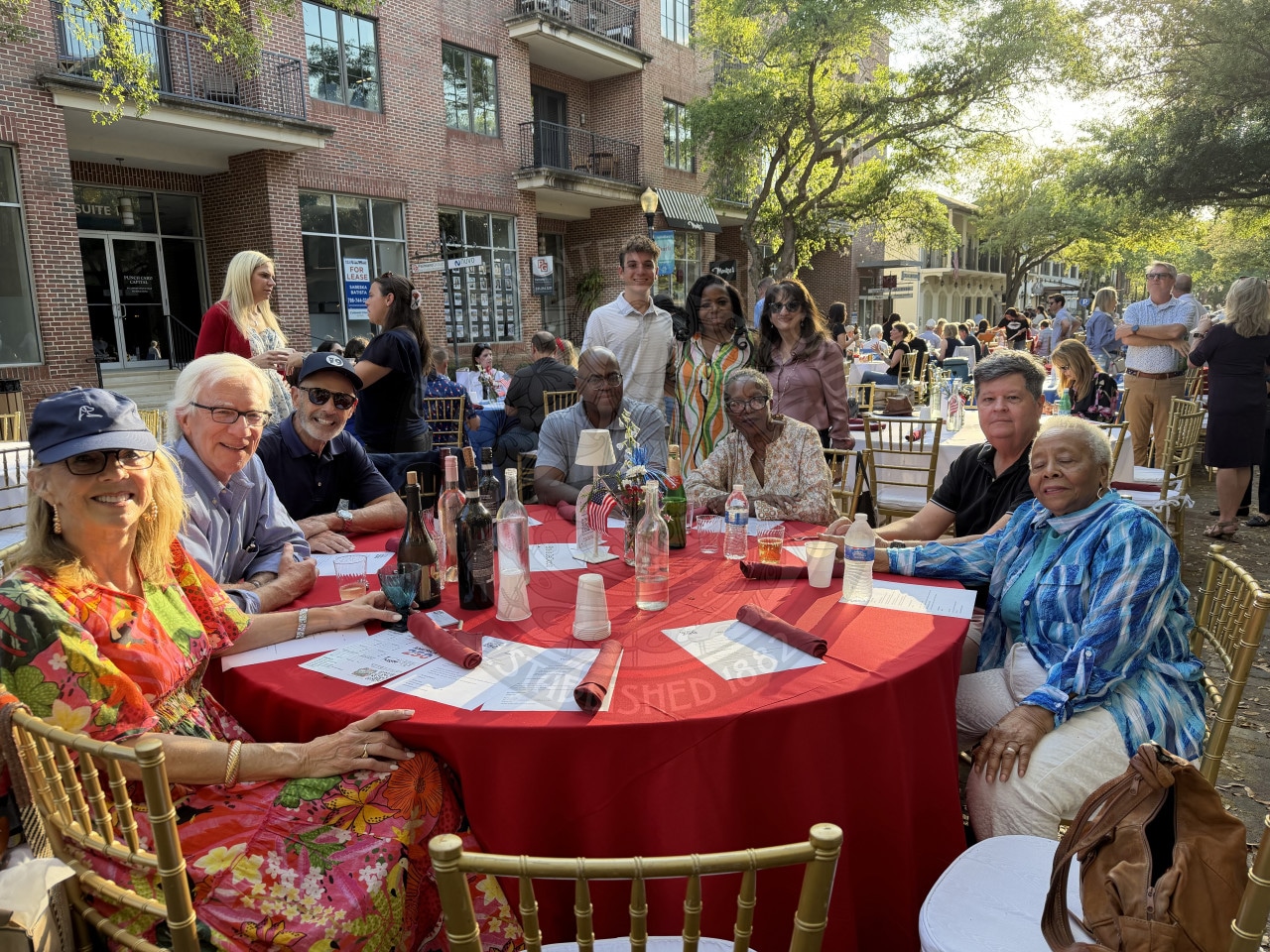 Seven people sit around a red table with three people standing behind them at an outdoors at a lively event, with drinks, menus, and buildings in the background.