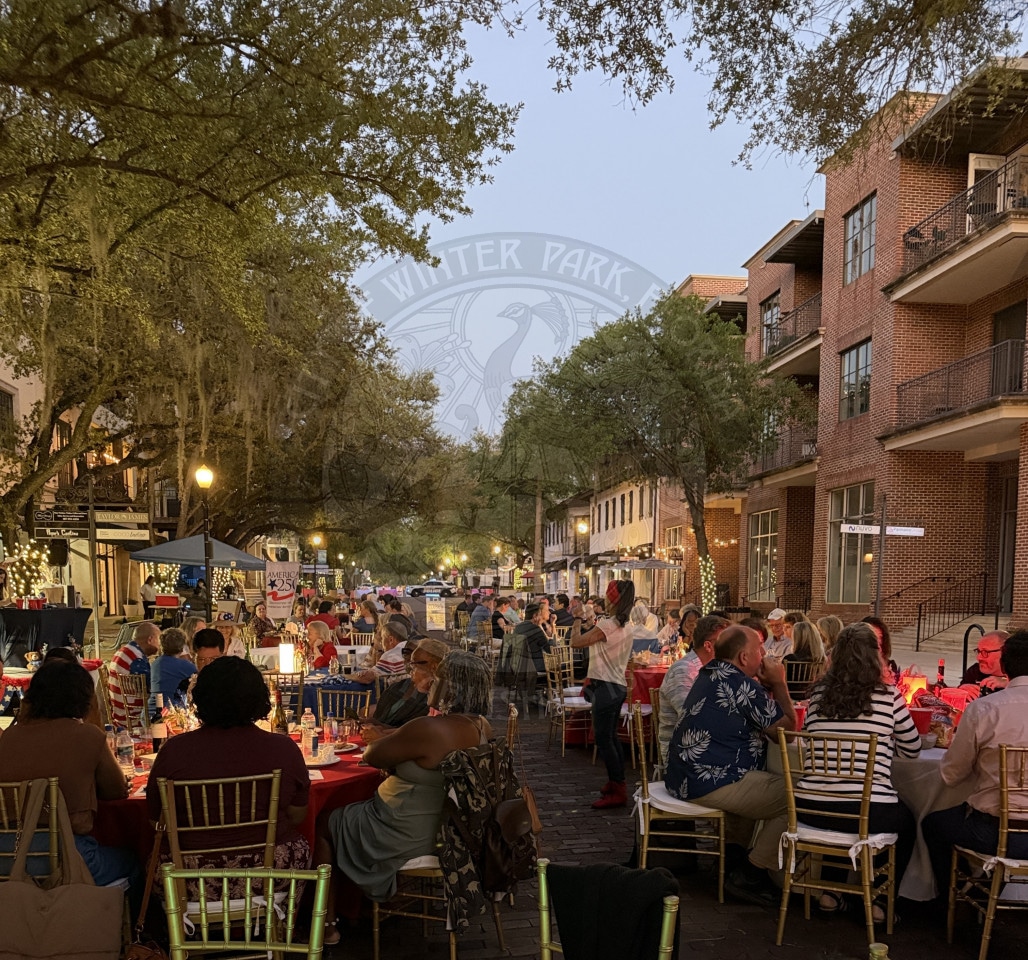 People dining at outdoor tables on a tree-lined street with string lights and brick buildings at dusk.