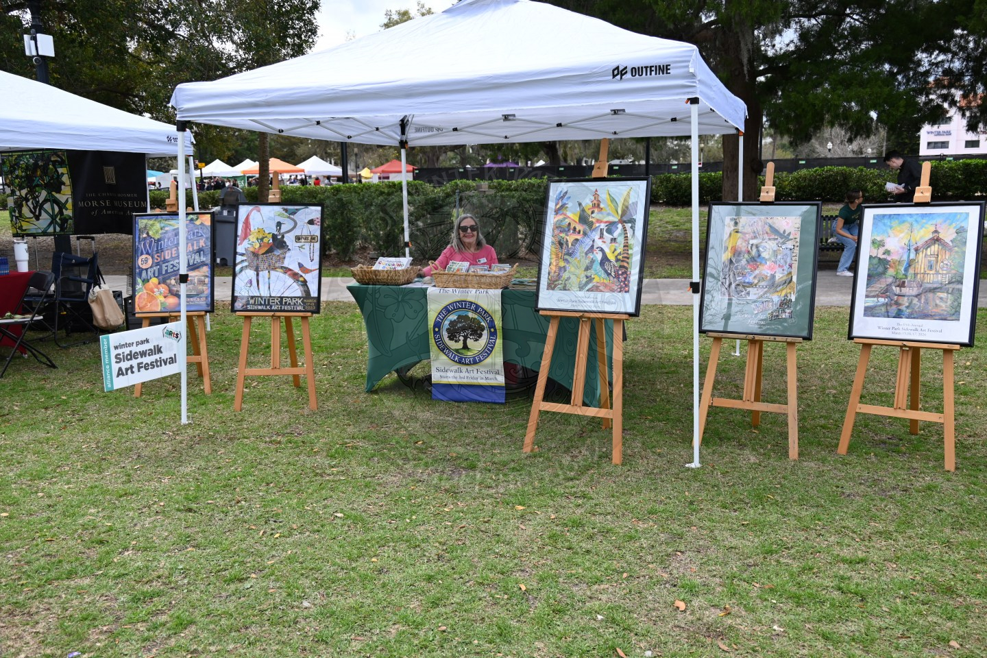 a woman sitting underneath tent behind a table with peices of art on easels in front of the tent.