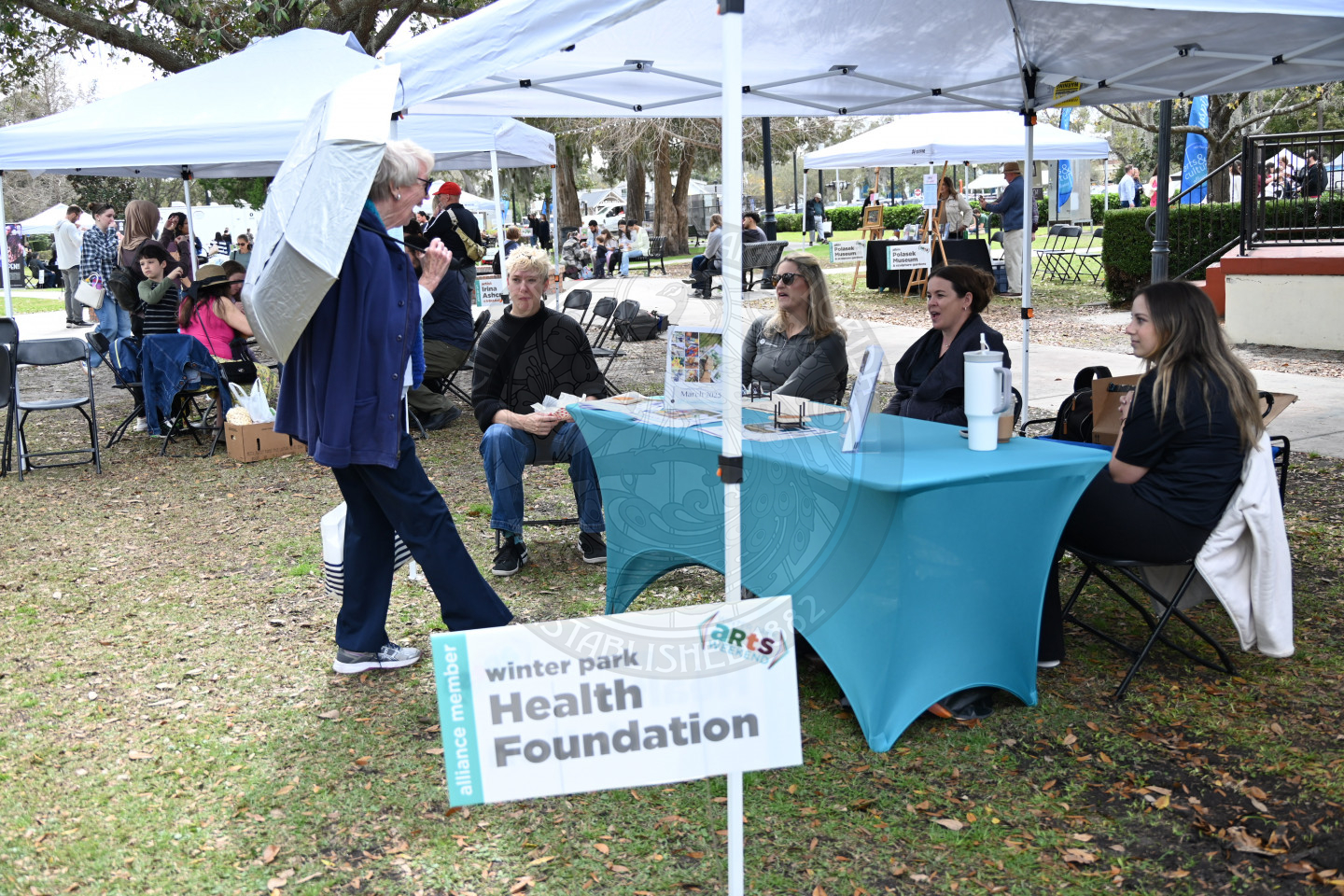 group of ladies sitting behind a table underneath a tent with an older lady holding an umbrella.