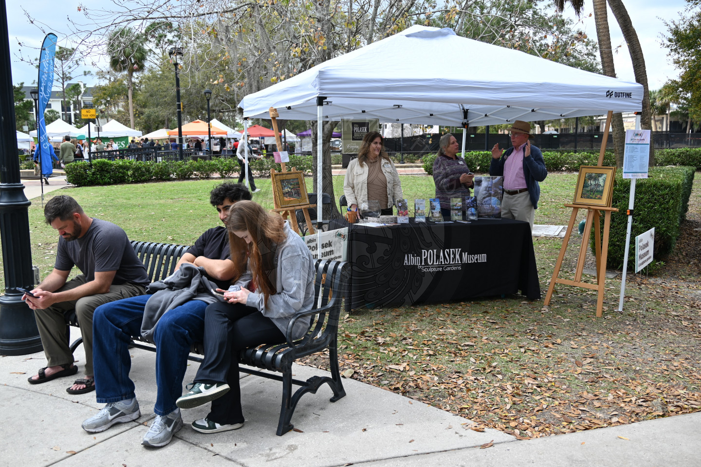 a park bench with people sitting on them and people standing underneath the tent behind a table that is under the tent.