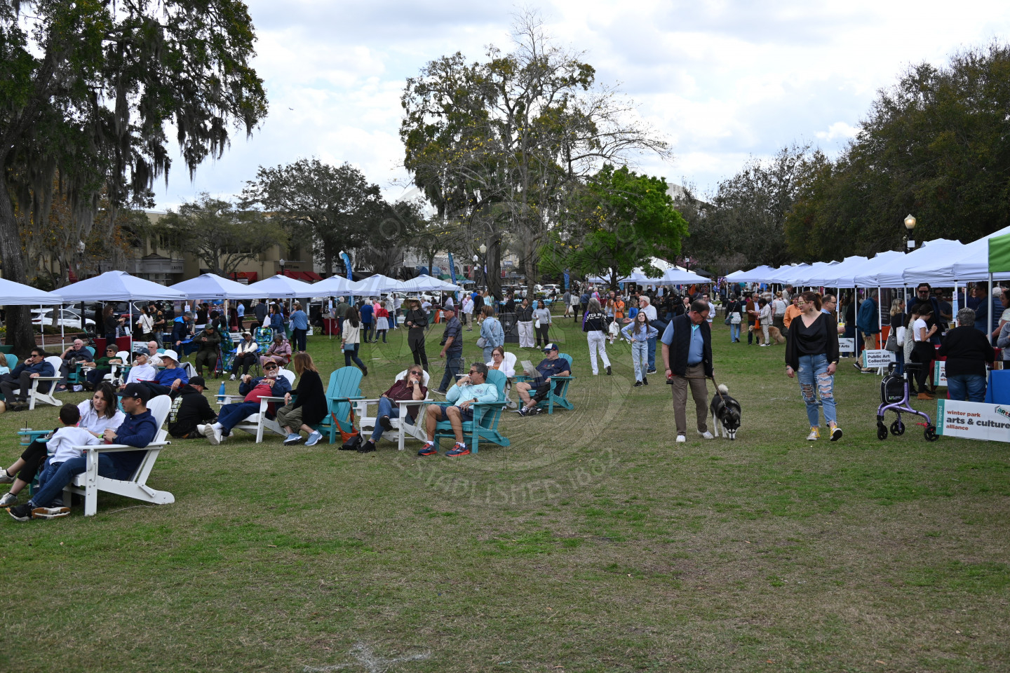 in a grass area people are sitting in chairs, standing and walking around browsing the tents along the sides of the grass.