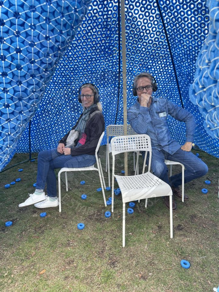 two people sitting in chairs in the blue element art peice with headphones on listening to the message that is playing.