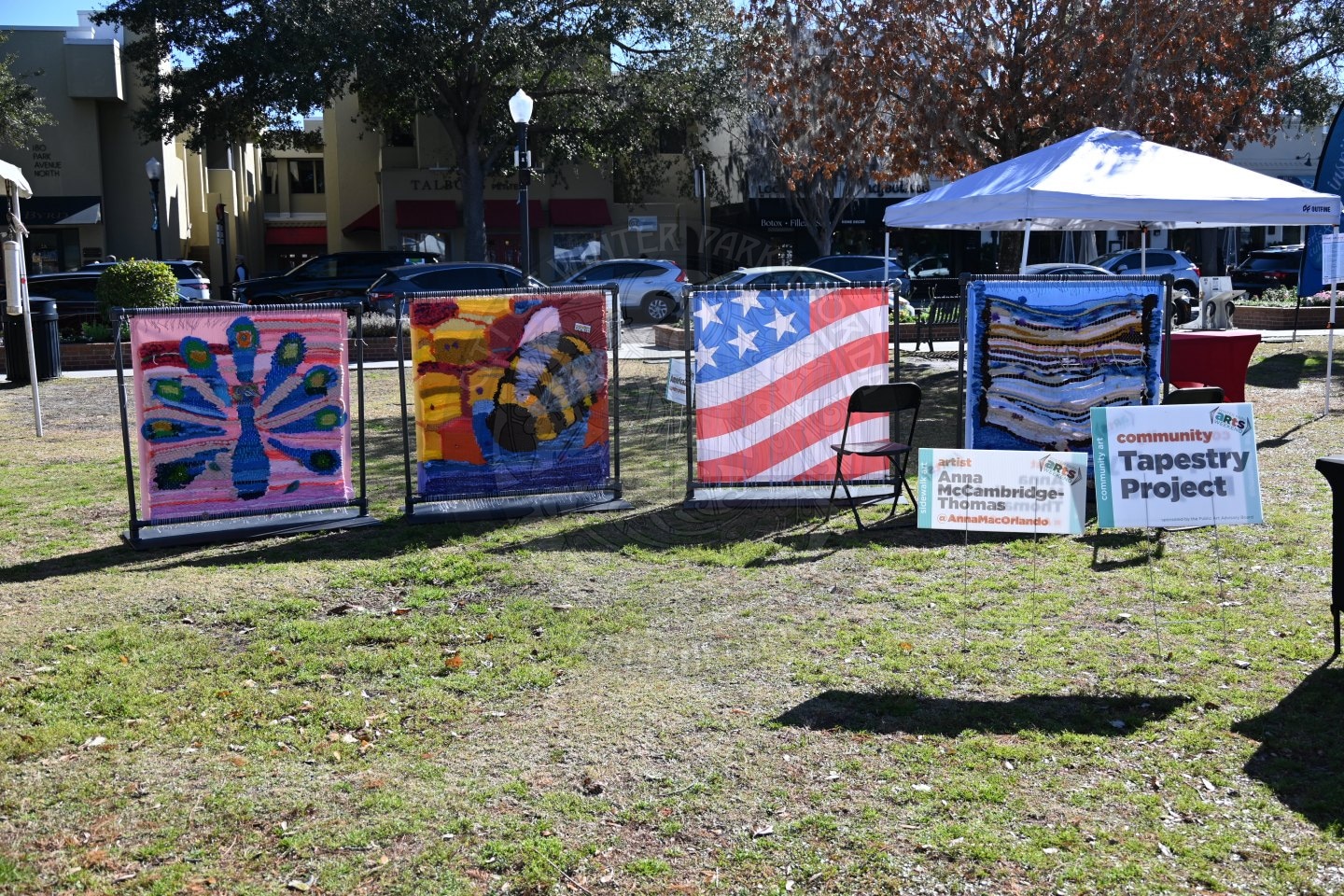 Four colorful tapestry panels, including a peacock and an American flag, are displayed outdoors on metal stands. A Community Tapestry Project sign is placed on the grass beside an empty chair.