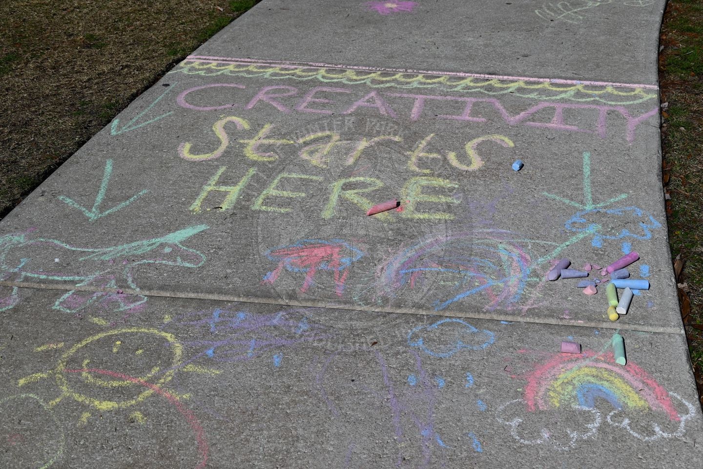Colorful sidewalk chalk art on pavement reads Creativity starts here with drawings of a unicorn, sun, rainbow, arrows, and scattered chalk pieces. The artwork is playful and vibrant.