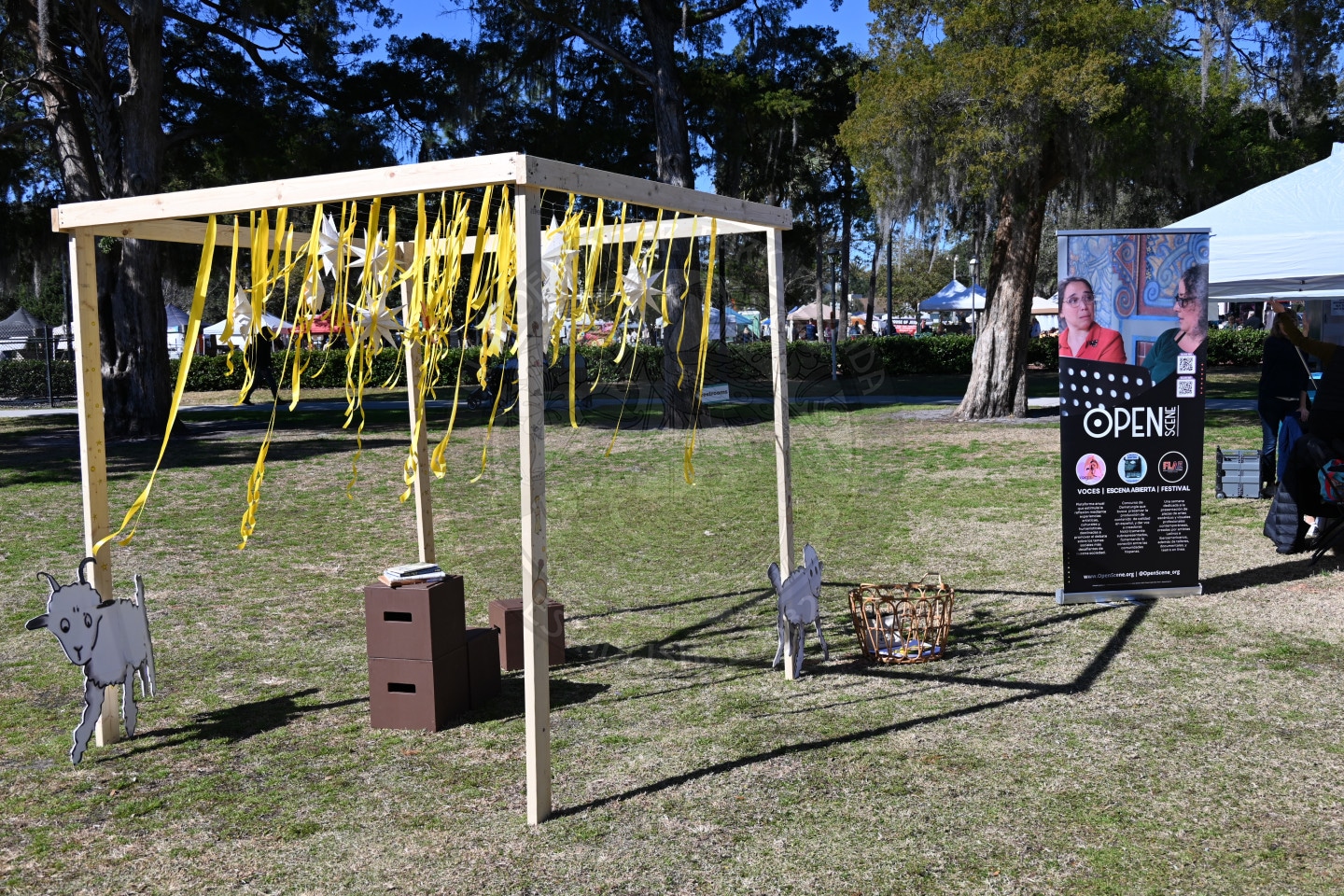 A wooden frame with yellow ribbons hanging down, animal cutouts, and boxes underneath stands on grass at an outdoor event. A large informational sign is to the right, with tents and trees in the background.