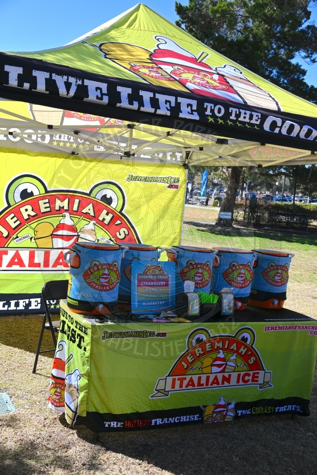 A Jeremiah’s Italian Ice stand at an outdoor event, featuring a bright green tent and tablecloth with the brand’s logo, colorful ice tubs, menus, and promotional signs displaying various flavors.