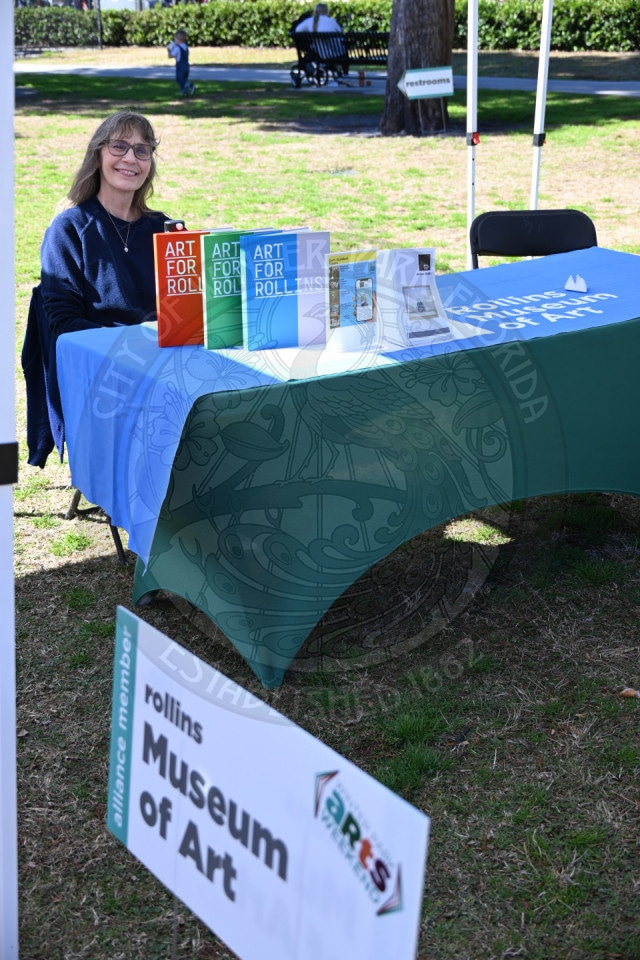 A smiling woman sits at an outdoor table covered with a green Rollins Museum of Art tablecloth, displaying colorful brochures. A museum sign stands in front on the grass.