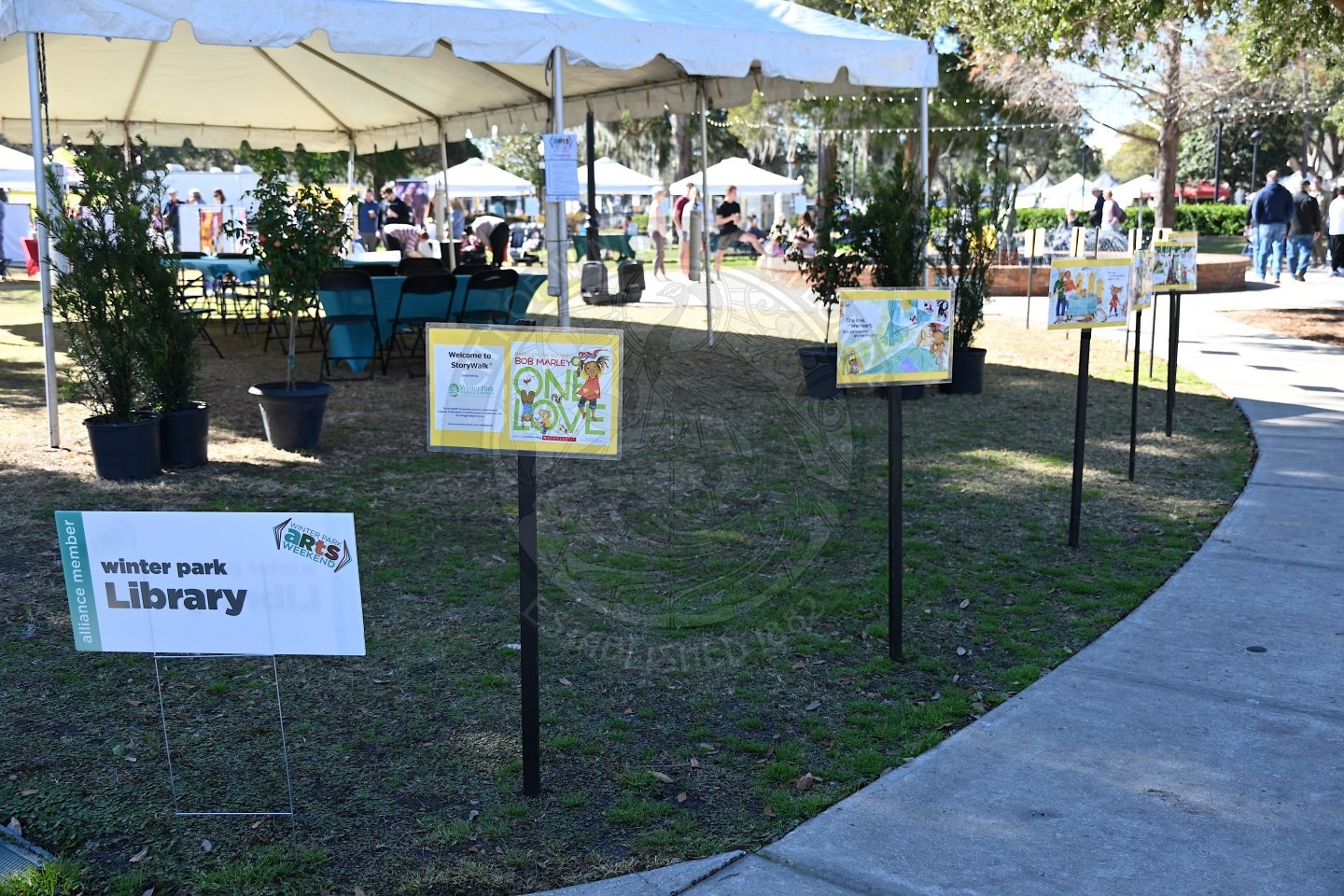 A grassy outdoor event features a row of illustrated storyboards on stands near a sign for Winter Park Library. A tent with tables and chairs is set up in the background under sunny skies.