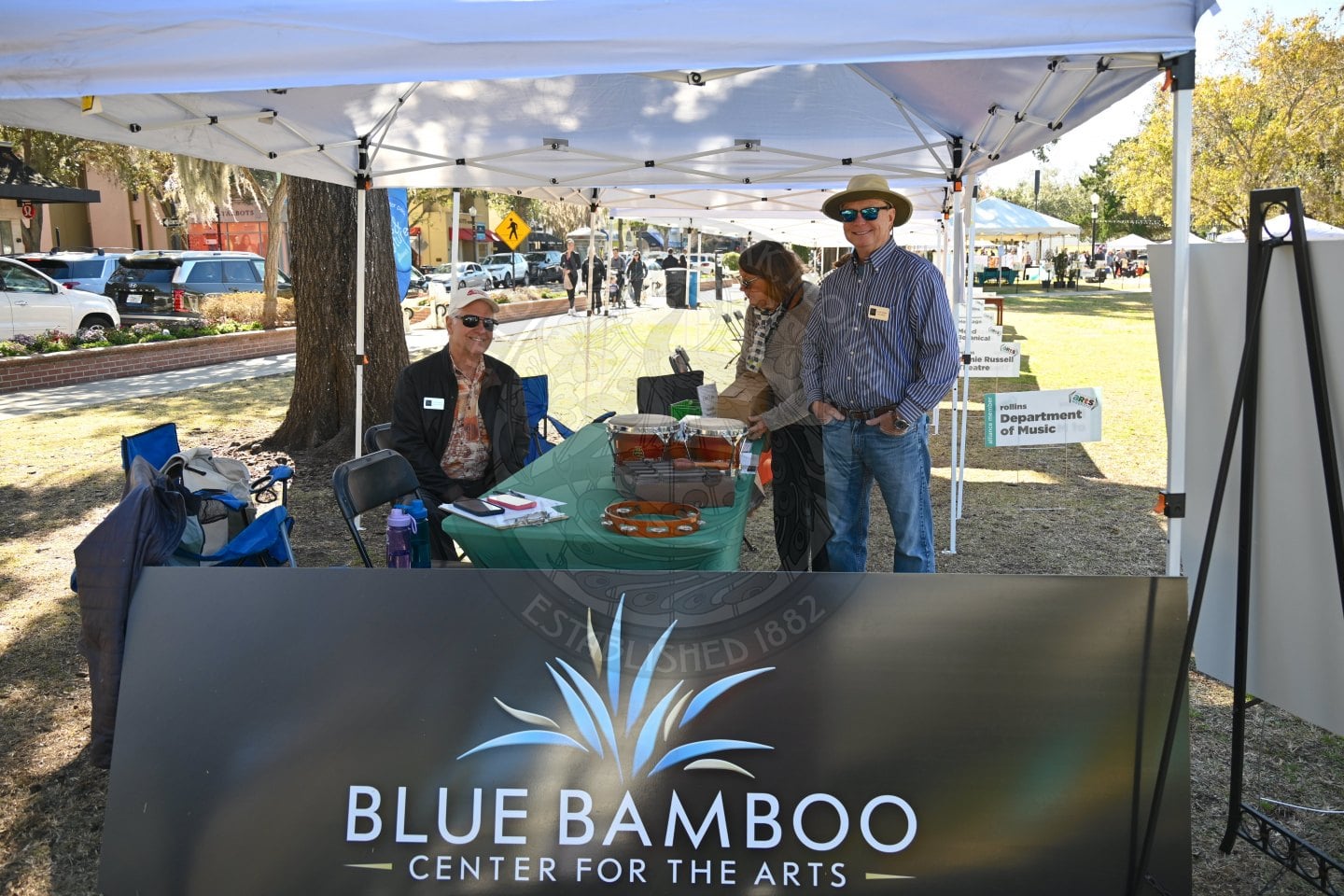 Three people stand and sit at an outdoor booth for the Blue Bamboo Center for the Arts. The booth is under a white canopy, with a large sign in front and trees, grass, and other tents in the background.