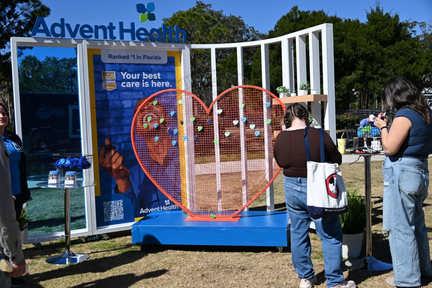 People stand near a heart-shaped display with notes attached, in front of an AdventHealth booth outdoors. A blue sign reads Your best care is here. Ranked #1 in Florida.