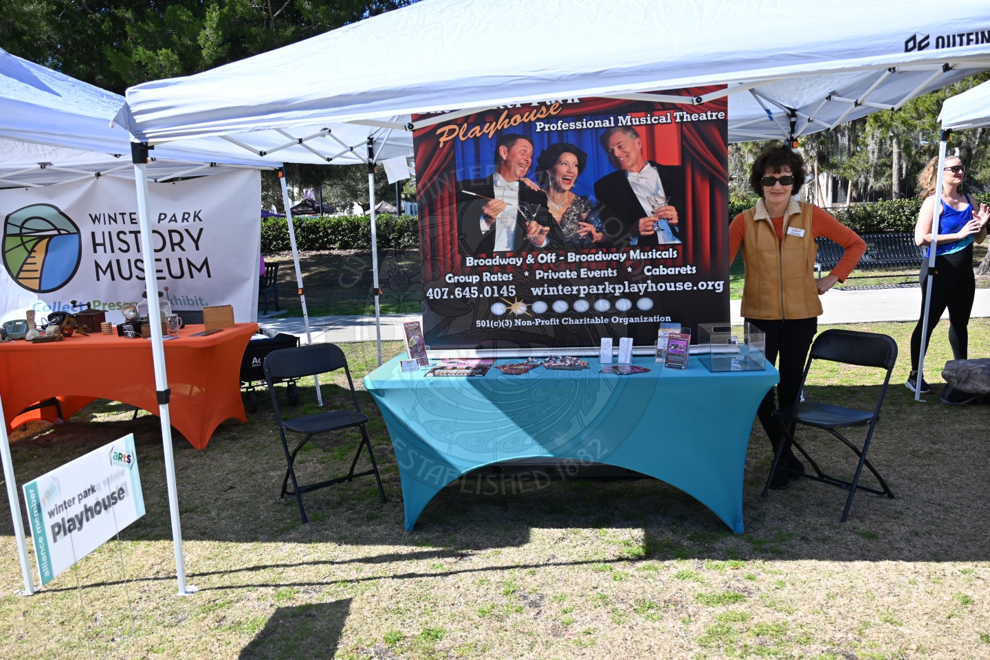 A woman stands and smiles next to a table under a tent at an outdoor event. The table displays flyers and a banner for the Winter Park Playhouse, advertising musical theatre, with another booth visible nearby.