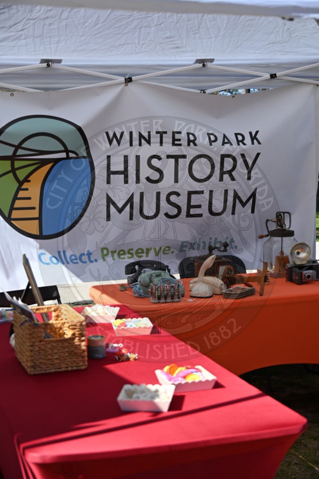 A booth with red tables displays vintage items and craft supplies in front of a banner that reads “Winter Park History Museum: Collect. Preserve. Exhibit.”.