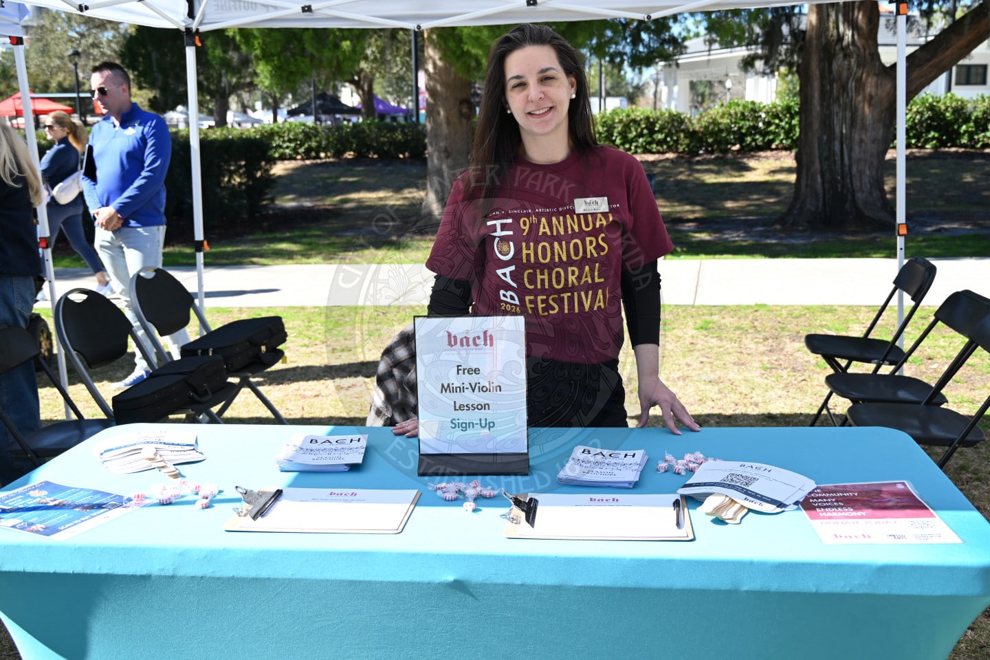 A woman stands behind a table with brochures, clipboards, and a Free Mini-Violin Lesson Sign-Up sign at an outdoor event under a canopy. She wears a maroon shirt and smiles at the camera.