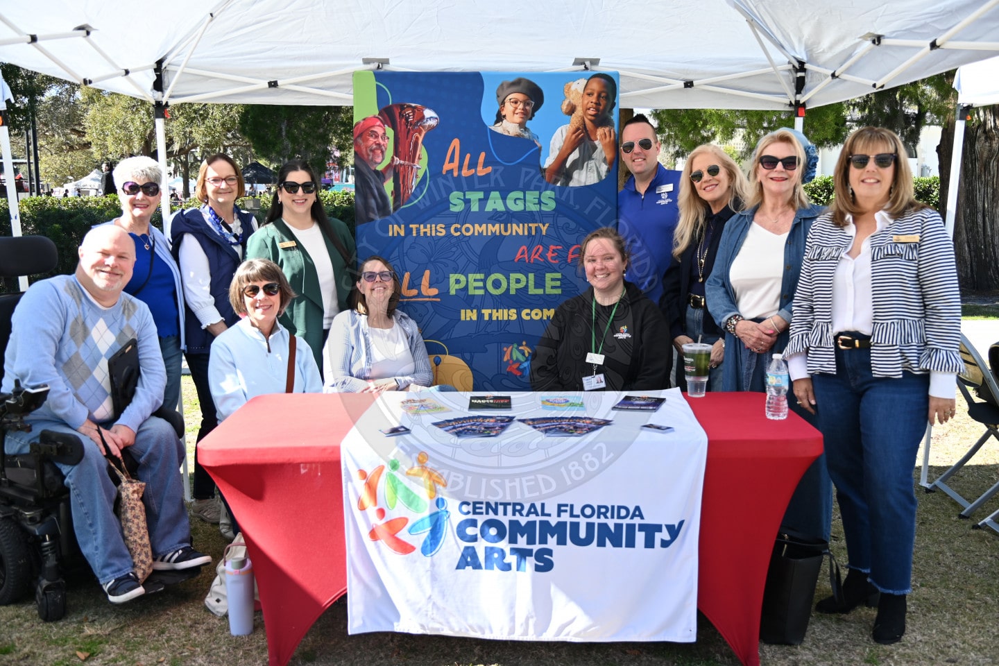 A group of people smiling and posing behind and around a table with a Central Florida Community Arts banner, under a tent, at an outdoor event. A colorful sign about inclusion is displayed behind them.