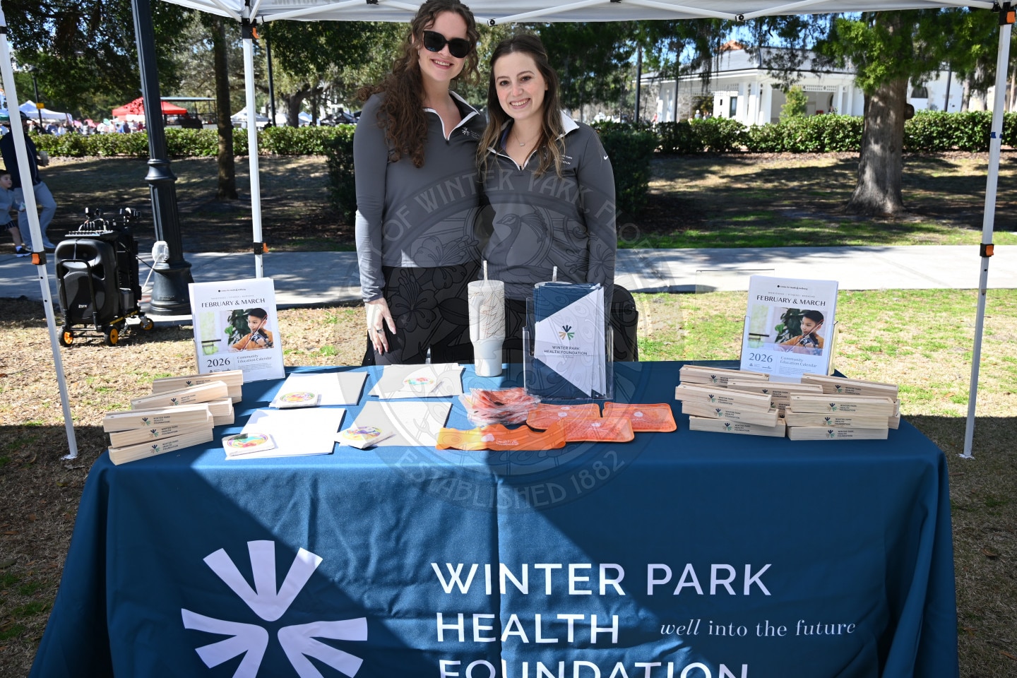 Two women stand behind a table covered with a Winter Park Health Foundation tablecloth, displaying brochures, boxes, and other health-related items at an outdoor event with trees and people in the background.