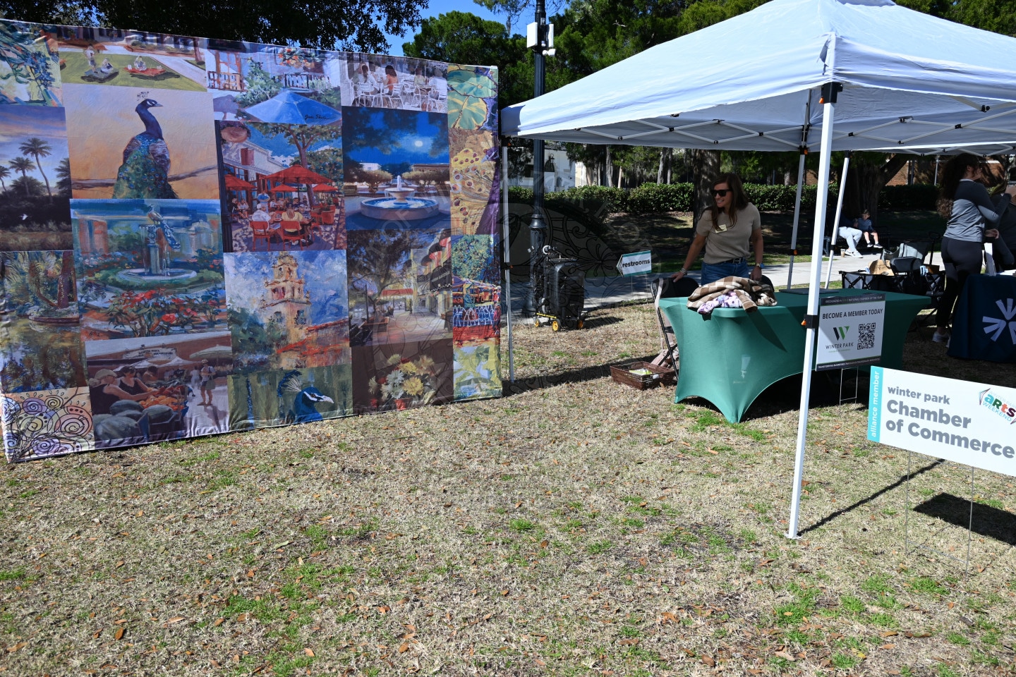 A colorful mural display with various paintings stands outdoors near a white tent, where a woman arranges items on a green table. A sign reads Winter Park Chamber of Commerce. Grass and trees are visible.