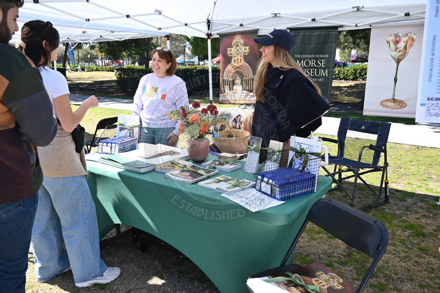 Two women stand behind a table covered with art prints, books, and flowers at an outdoor event booth, talking and smiling with two visitors. White tents and park scenery are visible in the background.