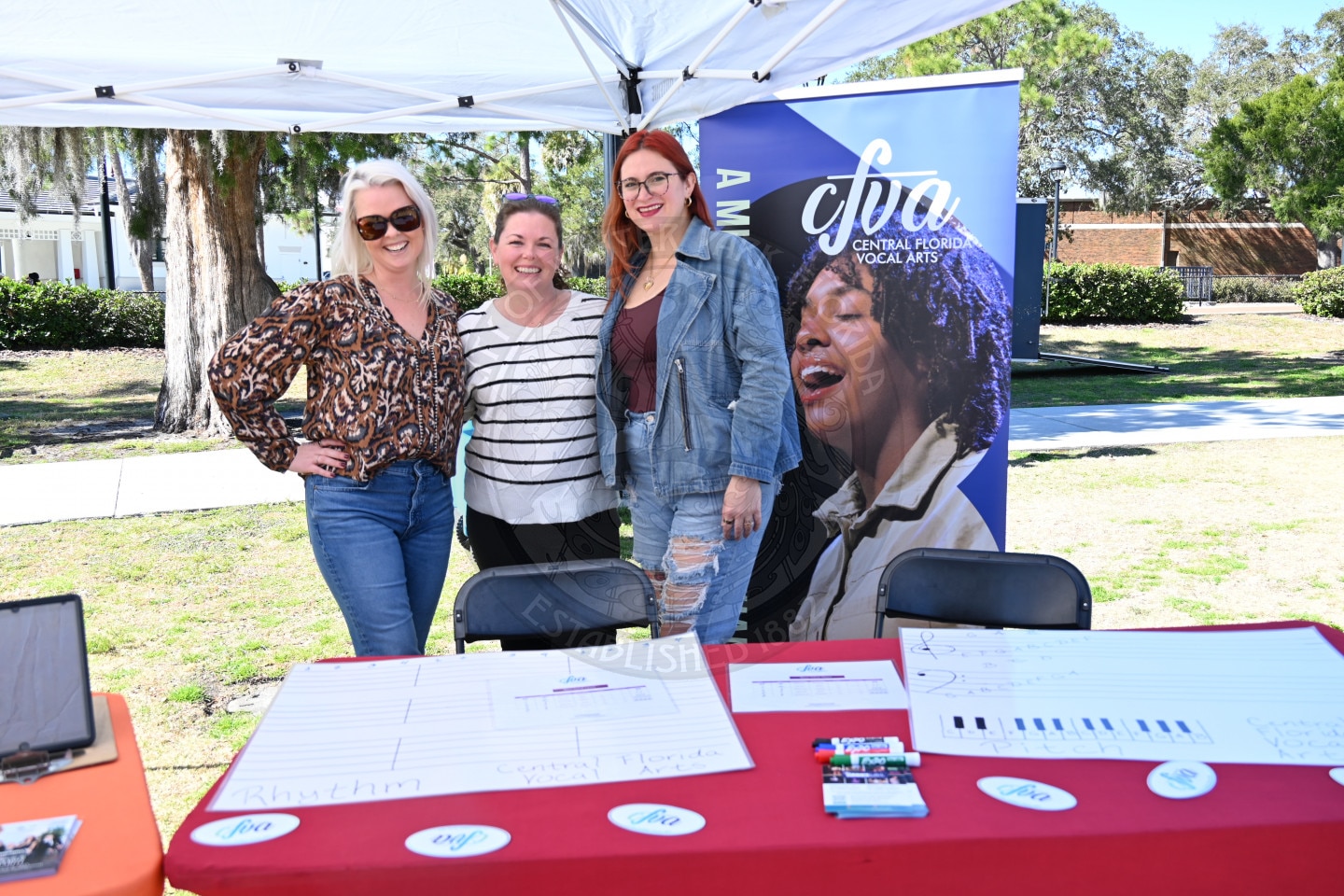 Three women stand and smile behind a table with music sheets, markers, and stickers at an outdoor event. A banner for the Central Florida Vocal Arts is displayed behind them, featuring a singer.