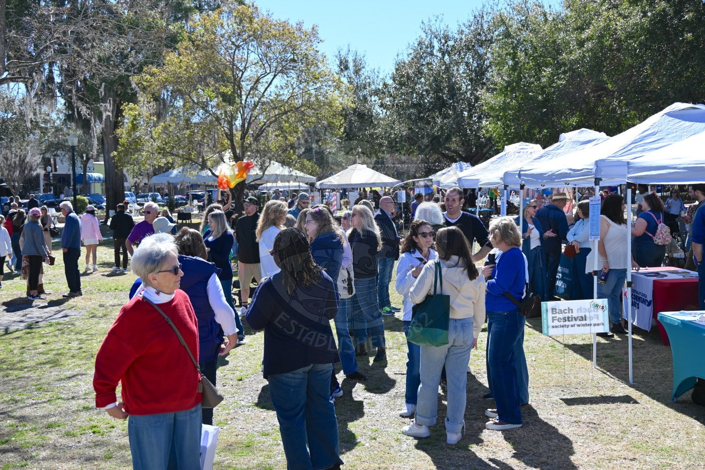 A crowd of people walks among white tents at an outdoor festival on a sunny day. Some people are talking, while others browse vendor booths, including one with a sign reading Bach Festival.