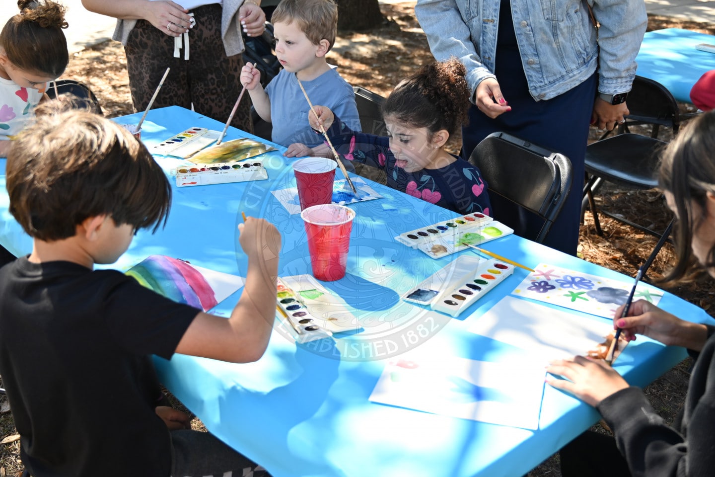 Children sit around a table covered with a blue tablecloth, painting with watercolors on white paper. Art supplies and red plastic cups are on the table, and adults supervise nearby.