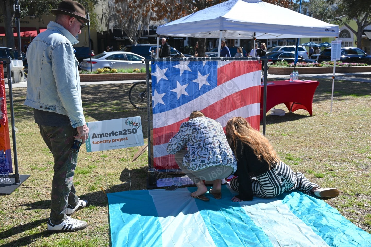 Three people work together on an art project featuring a large American flag at an outdoor event. One man stands watching the two others kneel by the flag.