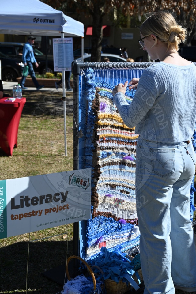 A woman stands outdoors, weaving colorful yarn into a large upright loom for a Literacy tapestry project. A sign and table are nearby, and other people and tents are visible in the background.