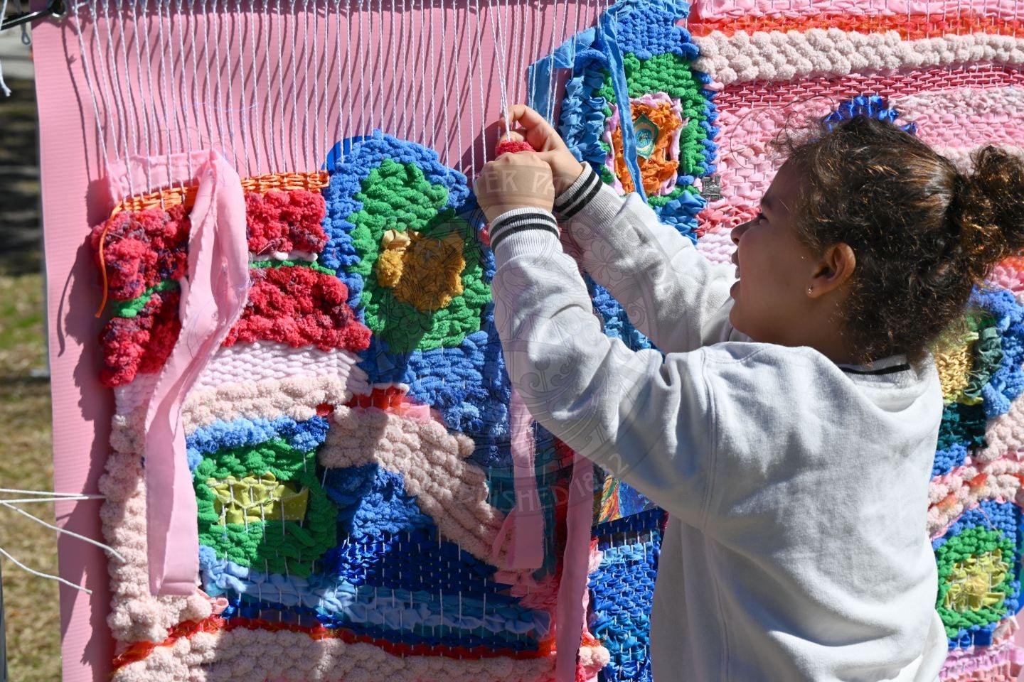A young girl weaves colorful yarn into a large, textured tapestry using a pink loom outdoors on a sunny day. She smiles and focuses on her creative work.