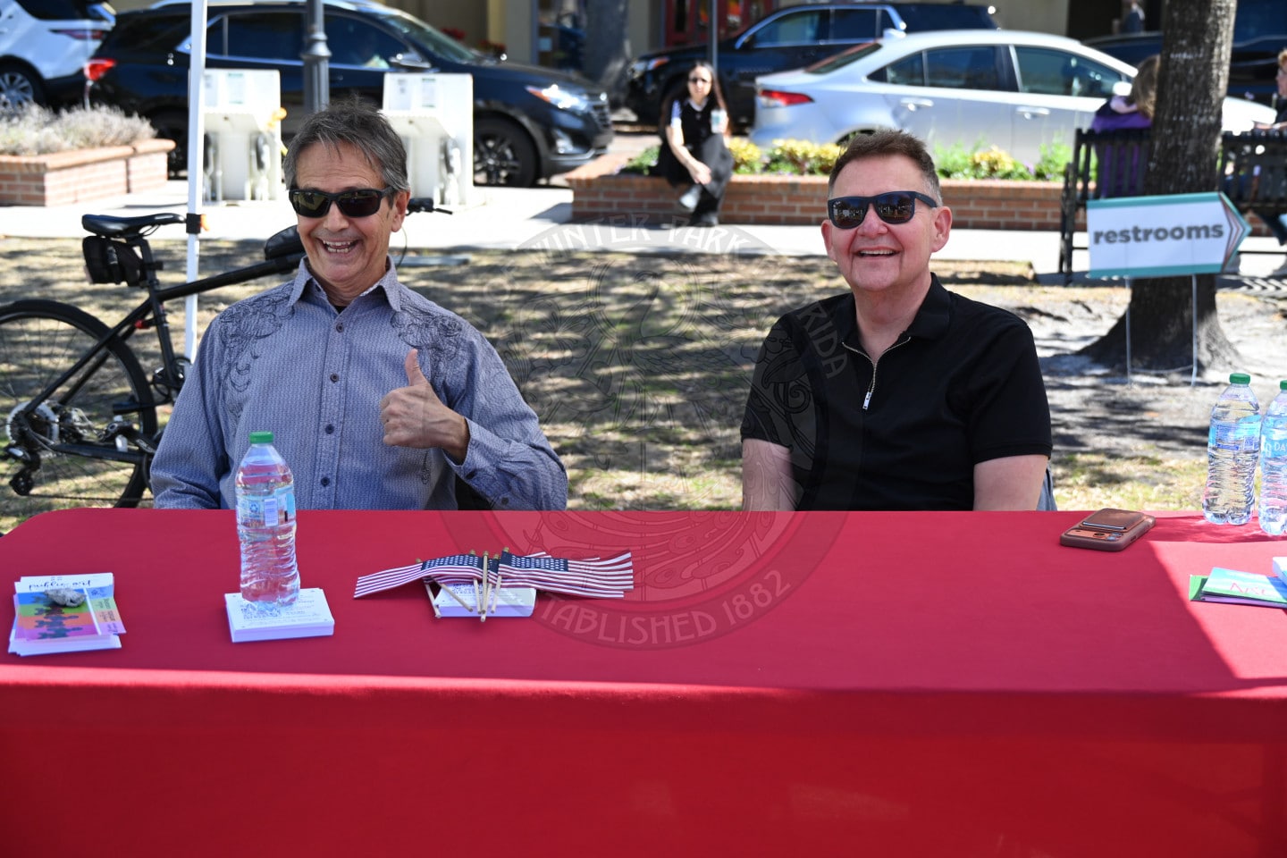 Two men sit at a red table outdoors, smiling. One gives a thumbs-up. Water bottles, pamphlets, and small US flags are on the table. Parked cars and a restrooms sign are visible in the background.