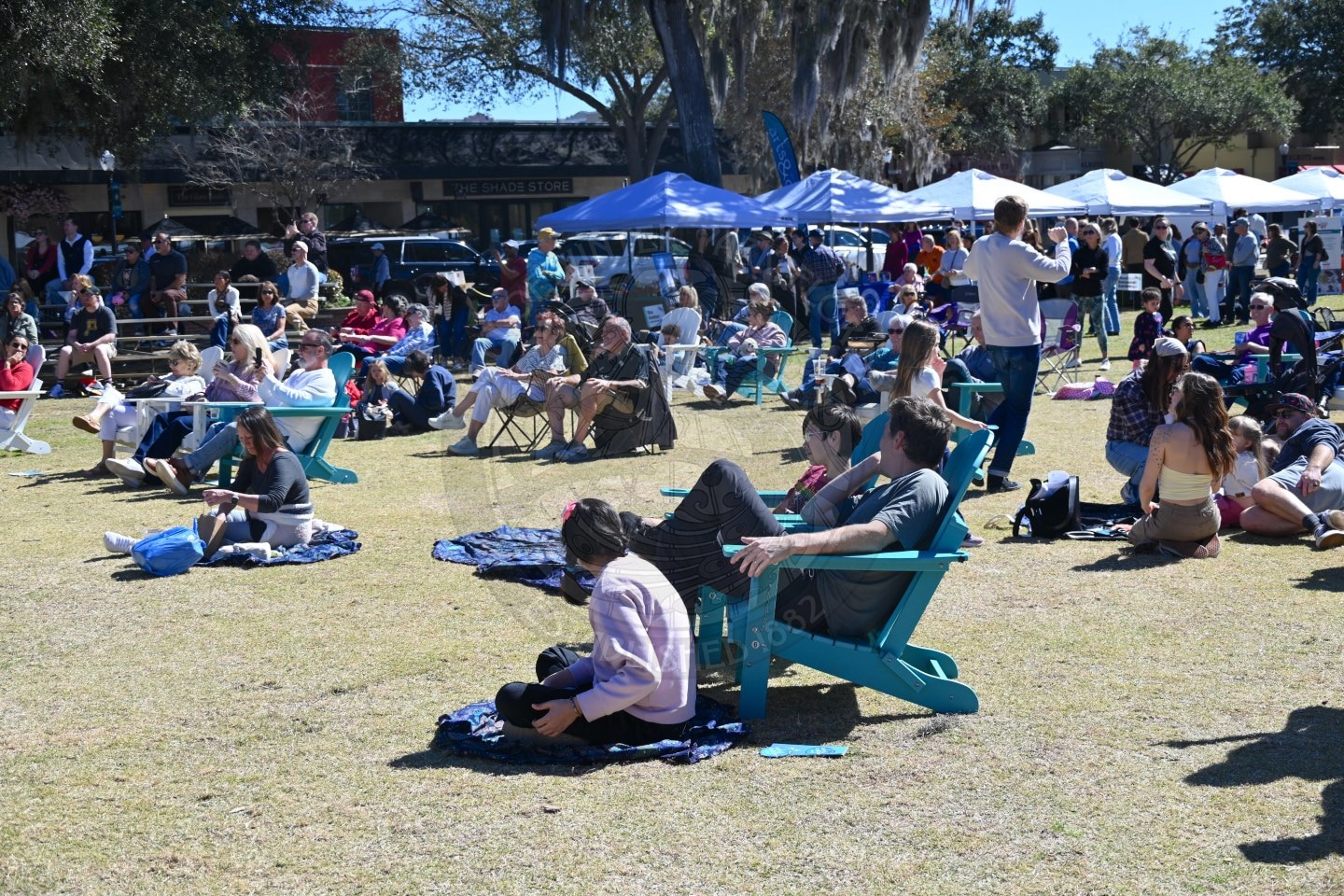 People sit on lawn chairs and blankets in a grassy park, watching an outdoor event. Tents and trees are visible in the background under a sunny sky. The crowd appears relaxed and casually dressed.