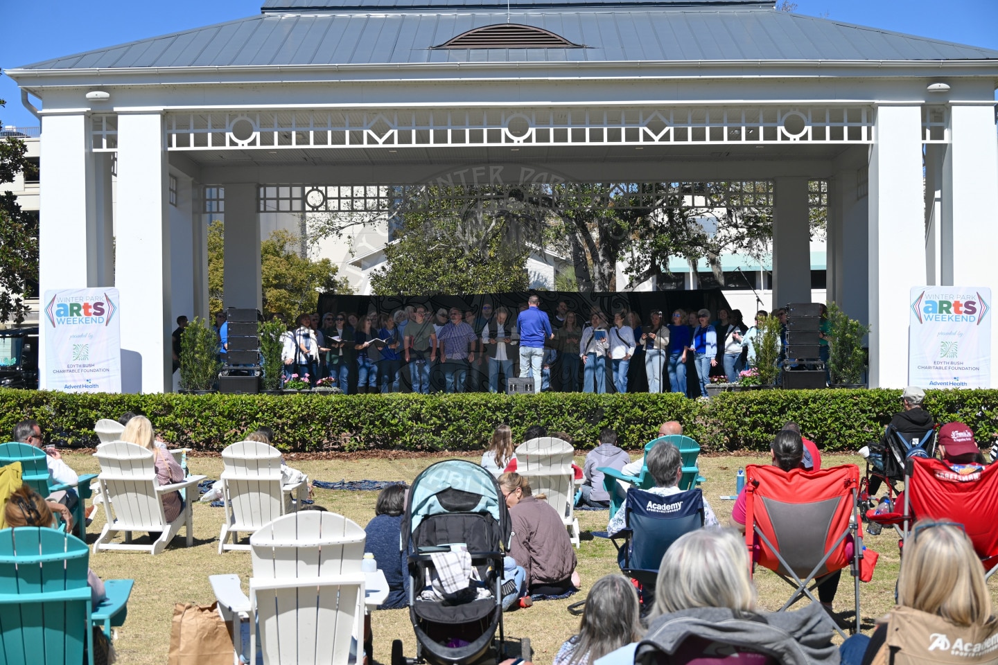 A group of people perform on an outdoor stage under a pavilion, while an audience sits on lawn chairs and blankets, enjoying the event on a sunny day. Arts Weekend banners are displayed on each side of the stage.