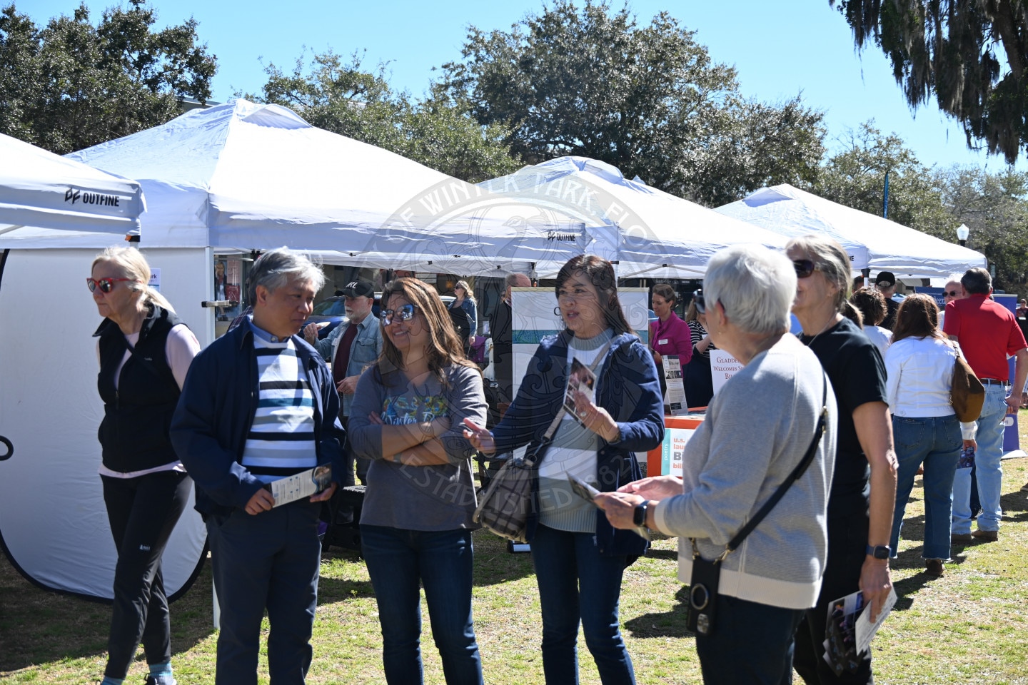 A group of people stand and talk in front of white tents at an outdoor event on a sunny day. Some hold pamphlets, and trees are visible in the background.
