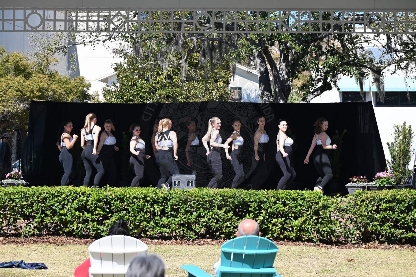 A group of young women in matching outfits perform a dance routine on an outdoor stage, while a few people watch from colorful chairs in the foreground. Trees and a building are visible in the background.