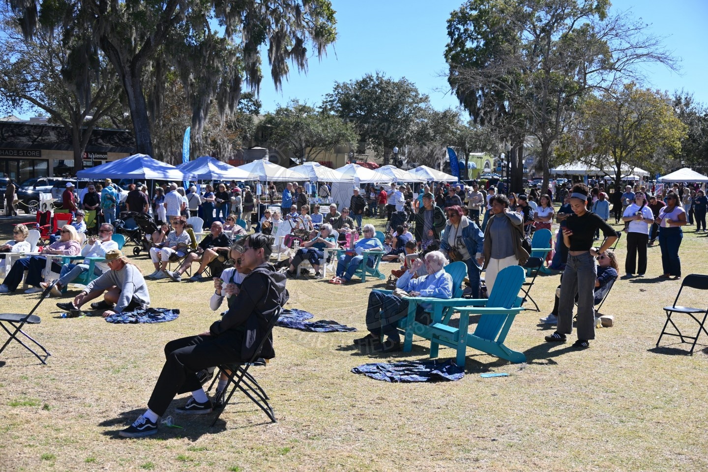 A large crowd of people sits on chairs, picnic blankets, and blue Adirondack chairs at an outdoor festival with tents and trees in the background on a sunny day.