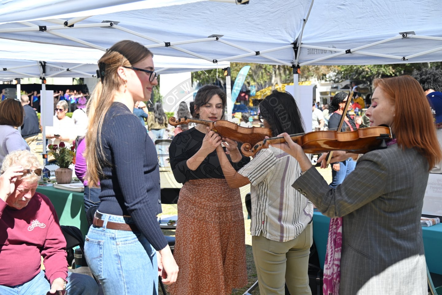 Three women stand under a white canopy at an outdoor event. Two are playing violins while the third watches. Other people and display tables are visible in the background.