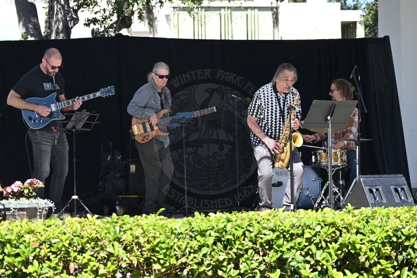 Four musicians perform outdoors on a stage: one plays electric guitar, another bass guitar, a third plays saxophone, and the fourth is on drums. A black backdrop and some plants are visible in the foreground.