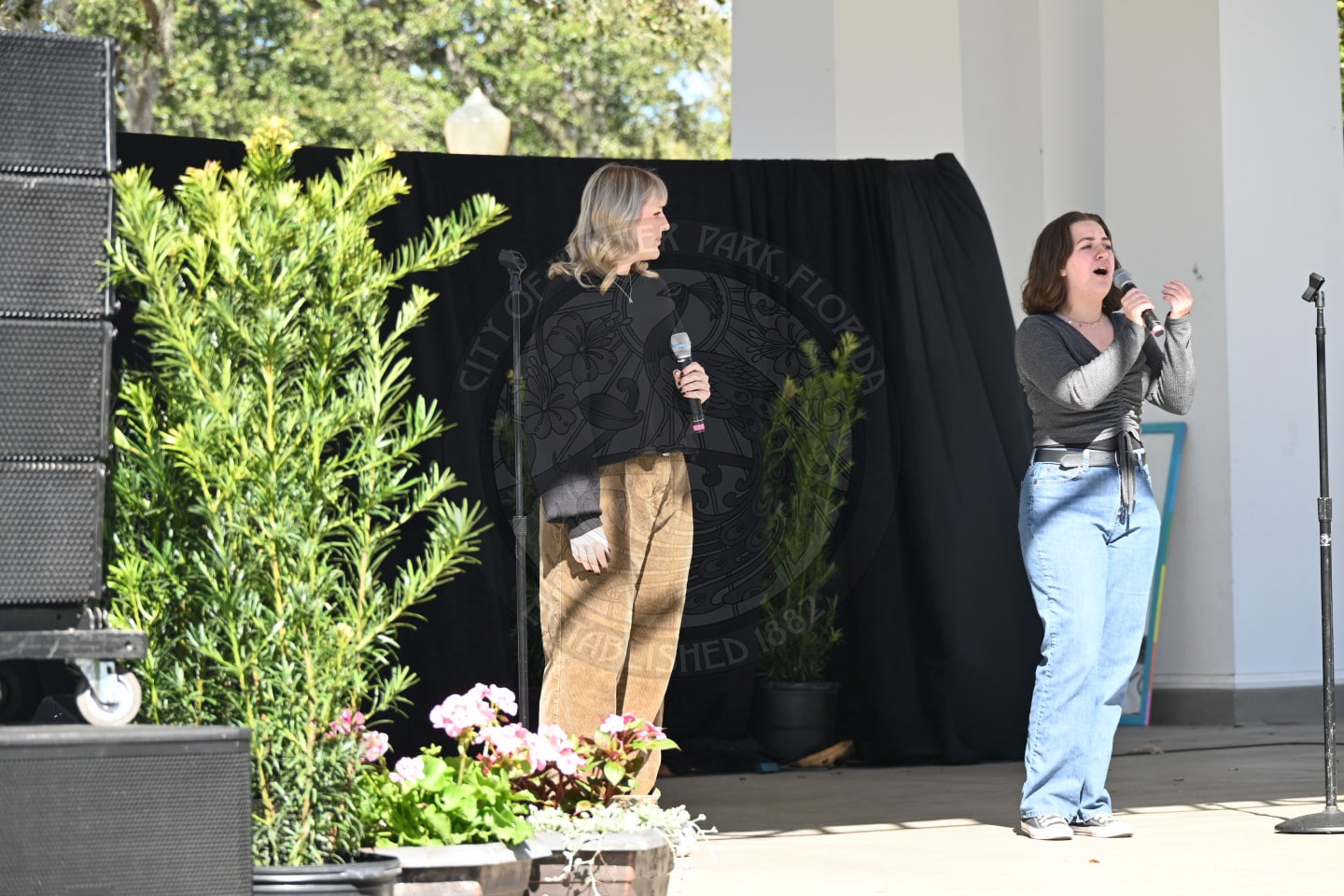 Two women stand on an outdoor stage with plants and black curtains; one holds a microphone and sings passionately while the other looks on, smiling. Sunlight illuminates the scene.