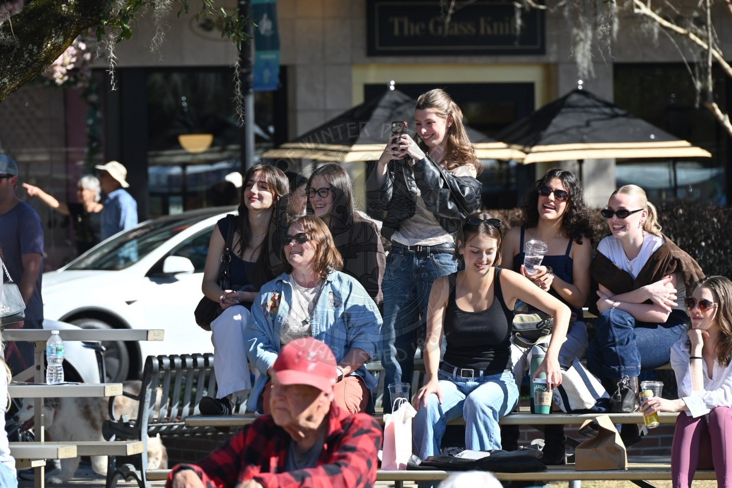 A group of smiling people sit on outdoor bleachers, some holding drinks. One woman stands to take a photo with her phone. Others chat and laugh together. Sunlight creates bright, cheerful atmosphere.