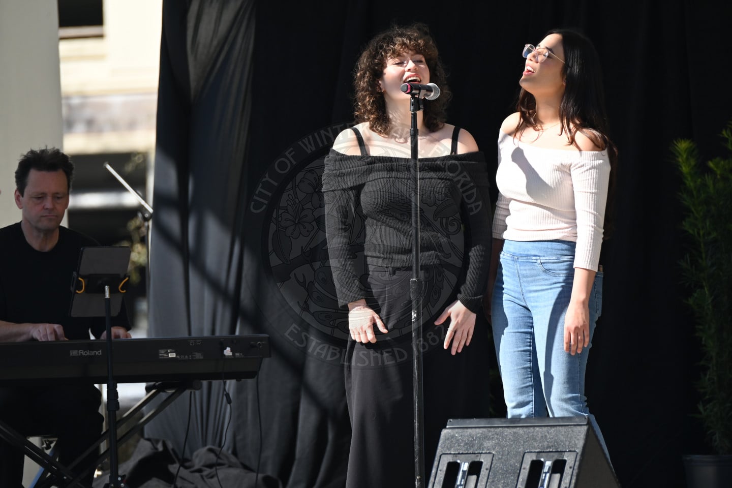 Two women sing together on stage, one at a microphone and the other smiling beside her. A man plays a keyboard to the left. They perform outdoors in front of a black curtain with a city emblem in the background.