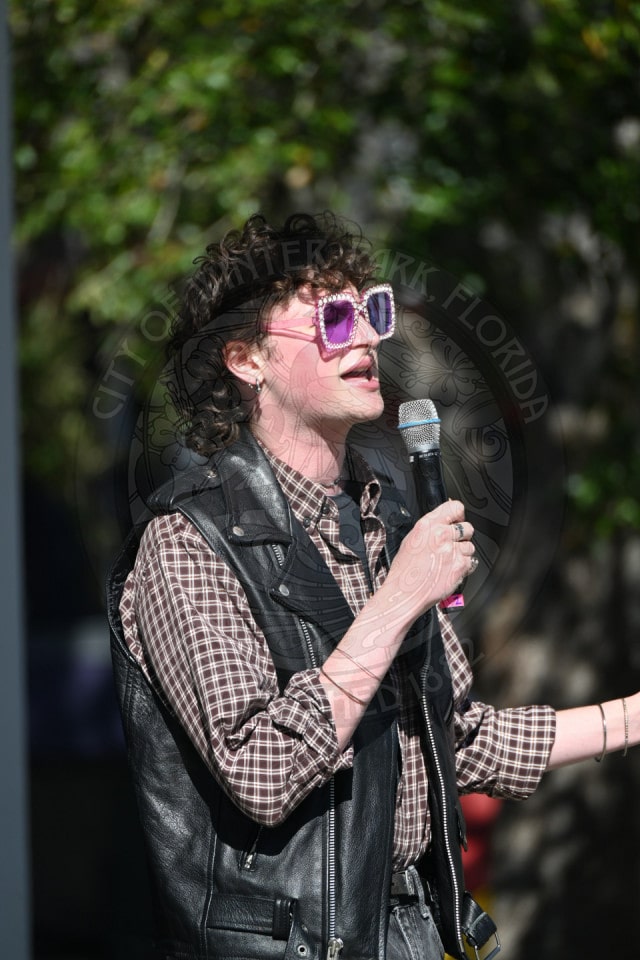 A person with curly hair, wearing pink sunglasses, a plaid shirt, and a black leather vest speaks into a microphone outdoors, with sunlight and greenery in the background.