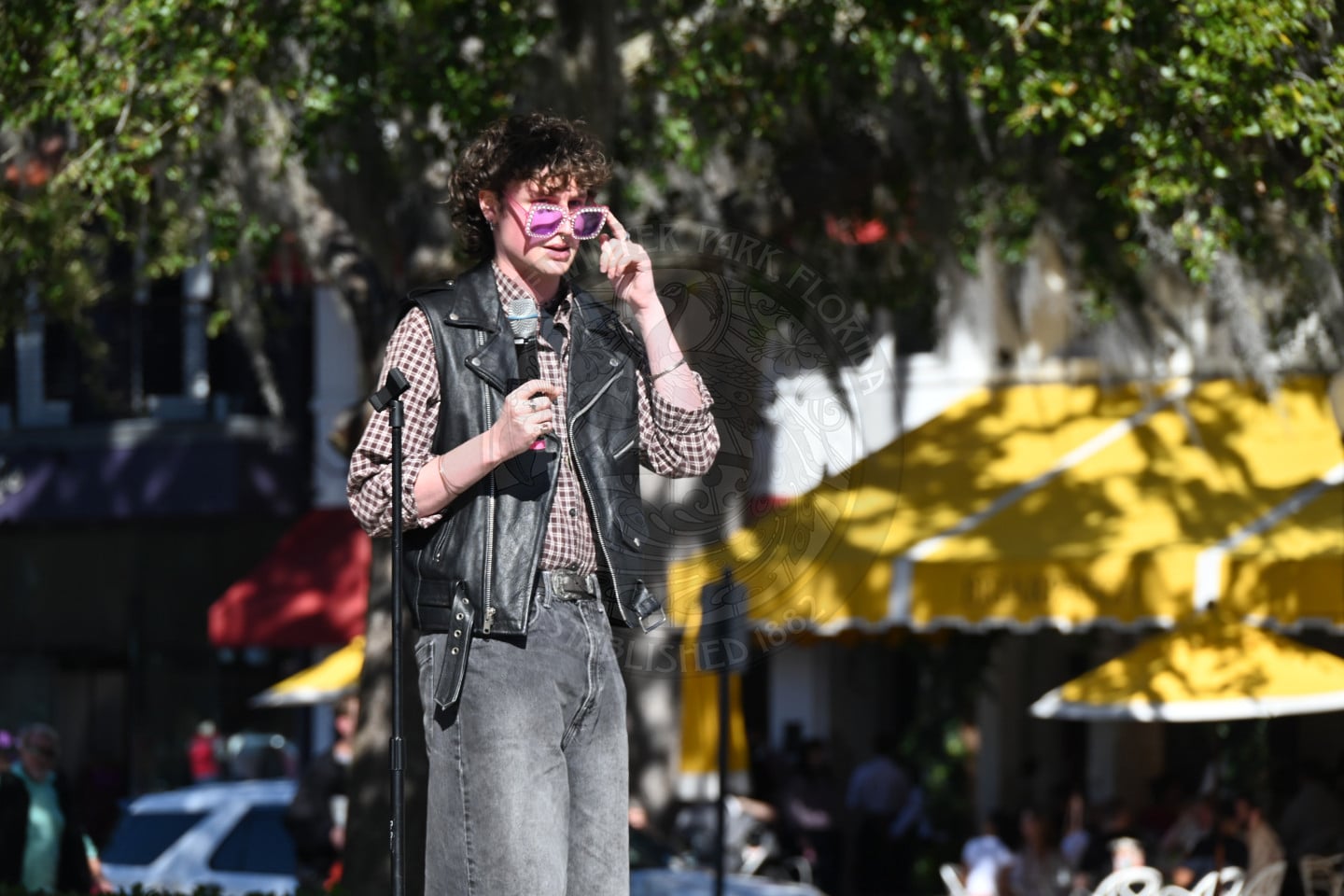 A person wearing pink sunglasses, a black leather vest, and a plaid shirt stands outdoors in front of a microphone. Behind them are yellow and red awnings, trees, and people seated at tables.