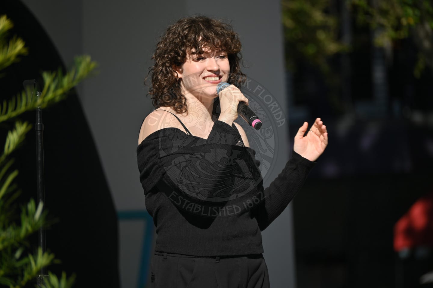 A person with curly brown hair, wearing a black off-shoulder top and pants, smiles while holding a microphone and performing outdoors in sunlight. Greenery and blurred background elements are visible.