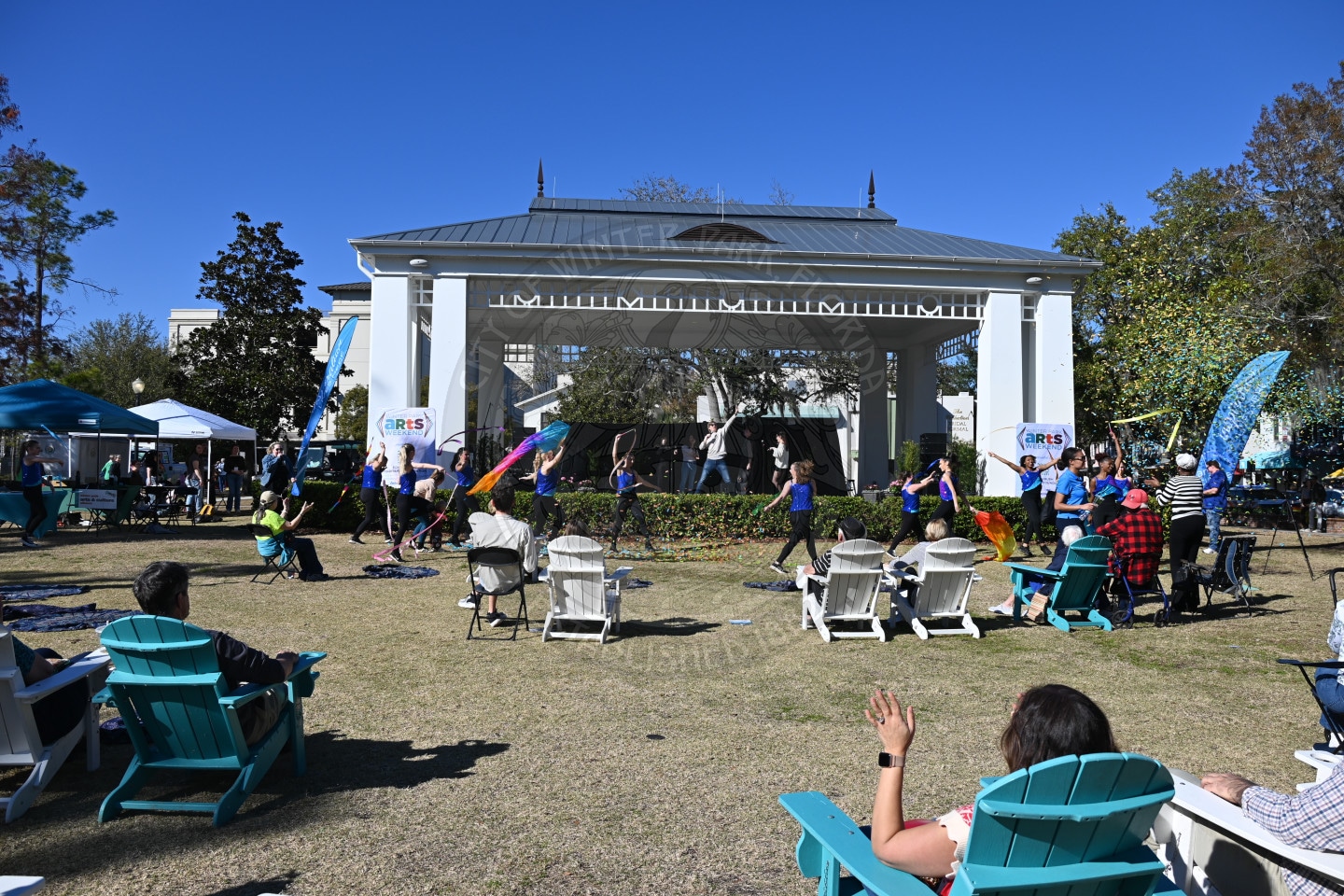 People watch a performance on an outdoor stage under a pavilion, with seated audience members in front. Flags and banners decorate the sunny scene, and trees surround the area.